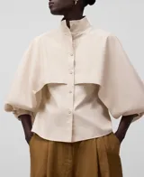 Camisa color crema de algodón, corte recto, con cuello mao y manga globo rematada en puño estrecho bajo el codo. Presenta botonadura frontal y capelina en la parte superior.
