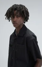 Camisa negra de manga corta estilo técnica, confeccionada en microfibra liviana. Tiene doble bolsillo frontal con cierre de cremallera y broches en lugar de botones.