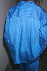 Camisa celeste con rayas verticales blancas, de corte oversize, cuello solapa, manga larga con puños y bolsillo de plastrón en el pecho.
