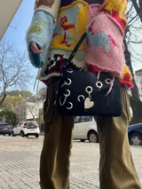 Cartera bandolera de cuero negro con ojalillos metálicos plateados, correa regulable con hebilla, bolsillo interior para labial y gancho para llaves. Diseño uruguayo.