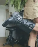 Bota alta de cuero vacuno color negro, con caña recta, puntera cuadrada y detalle de arnés con hebilla metálica plateada.