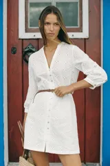 Vestido corto blanco con bordados calados, cuello redondo con escote en pico, manga tres cuartos, cierre frontal con botones y cinturón fino marrón.