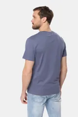 Remera blanca de manga corta con cuello redondo y un estampado gráfico frontal que simula un paisaje costero con un sol naciente o poniente, rodeado por texto circular que dice "SURF - LIFE UNDER THE SUN - CLUB".