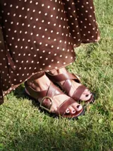 Sandalias bajas de cuero color marrón con tiras anchas cruzadas sobre el empeine y hebilla metálica dorada en forma de estribo.