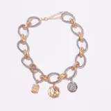 Collar de eslabones gruesos entrelazados en tonos plateado y dorado, con tres dijes colgantes: uno rectangular dorado con relieve de rostro, uno redondo dorado con textura martillada y uno redondo plateado con diseño floral o mandala.