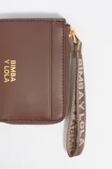 Monedero pequeño de estructura rígida, color negro, con cierre de cremallera y tirador de tejido amarillo mostaza. Presenta un bolsillo exterior frontal y el logo de la marca grabado en dorado.