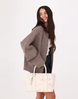 Cartera tipo tote color blanco crudo, con doble asa corta y correa larga regulable y desmontable. Incluye charm colgante con forma de corazón y cierre superior.