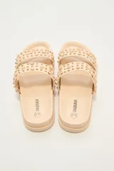 Sandalia tipo slide de color crudo, con dos tiras anchas adornadas con tachas doradas redondas de diferentes tamaños. La suela es gruesa y de goma.
