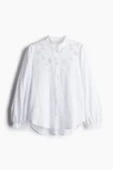 Blusa blanca de corte holgado en tejido liviano de lino y viscosa con canesú delantero en broderie. Cuello mao con frunces en la nuca, botones forrados adelante y mangas globo largas con puños finos abotonados. Bajo redondeado.