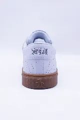 Zapatilla urbana de caña baja, color blanco, con cordones blancos y suela de goma color caramelo. Presenta microperforaciones en el lateral y el logo de un pingüino bordado.