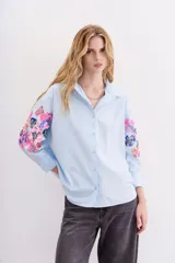 Camisa blanca de algodón con bordados florales y lentejuelas en las mangas, cuello camisero, mangas largas con puños abotonados y corte amplio.
