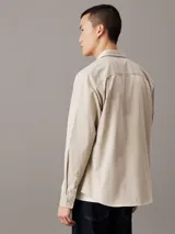 Camisa de manga larga color beige claro, con cuello clásico, cierre frontal con botones y logo de la marca bordado en el pecho.