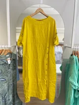 Vestido midi amarillo de lino, con corte recto, cuello redondo y mangas cortas con botón. Presenta una hilera de botones decorativos en el lateral.