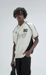 Chomba tejida de punto cerrado color off-white, con cuello polo y ribetes negros. Presenta un diseño deportivo con paneles laterales negros, número "90" y texto "Ravers" bordado en el frente.