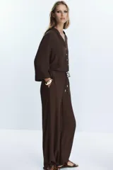 Pantalón ancho de tiro alto color marrón oscuro, con cintura elástica ajustable mediante cordones con terminales doradas. Presenta textura arrugada en la tela y bolsillos laterales.