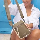 Bolso pequeño tipo bandolera o phone bag, de material símil cuero, predominantemente color mostaza/amarillo, con detalles en marrón oscuro en los bordes y un panel lateral color beige claro. Posee correa ancha de tela regulable y desmontable, y un bolsillo frontal con cierre.