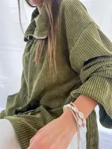 Buzo de pana fina color verde oliva, con diseño de corte holgado y mangas largas. Presenta una textura de cordón vertical característica de la pana y un pequeño parche decorativo en la parte superior de la espalda.