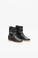 Bota de caña media color negro confeccionada en cuero, con suela de goma y terminaciones de costura manual y remaches metálicos. Presenta un cinto ornamental con tachas, cadenas y piezas en metal.