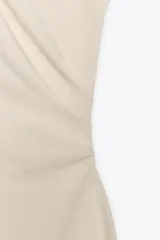Vestido midi color beige confeccionado en hilatura con algodón. Tiene cuello redondo, manga corta caída y detalle de drapeado lateral.