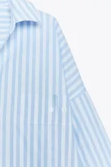 Camisa celeste y blanca a rayas verticales, de corte amplio, con cuello solapa, escote pico, manga larga con puños abotonados, bolsillo en el pecho y bajo asimétrico con aberturas laterales. Cierre frontal con botones.