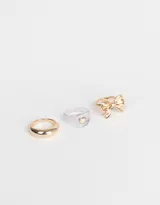 Set de tres anillos dorados: uno liso, uno transparente con un corazón dorado y uno con forma de moño.