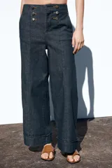 Pantalón culotte de tiro alto en denim azul oscuro, con pernera ancha y corta terminada en vuelta. Presenta falsos botones dorados en la parte delantera, bolsillos frontales y cierre con cremallera y gancho.