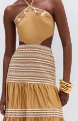 Vestido largo color ocre sin mangas, confeccionado en tricolina con forro. Presenta bordados en madera, hilo y pedrería, abertura en la cintura y tira para atar al cuello.