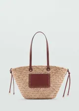 Bolso de mano tipo shopper de paja color beige con aplique rectangular de cuero marrón con tachas doradas. Tiene doble asa de cuero marrón y cierre ajustable con cordones.