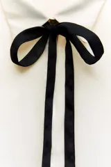 Camisa color crema de satén con cuello solapa, manga larga y lazo negro contrastante. Cierre frontal con botones.