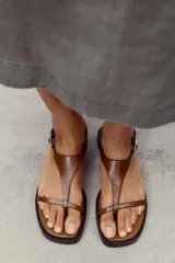 Sandalias planas color marrón con base negra, tira delantera fina y pulsera al tobillo con hebilla metálica dorada.