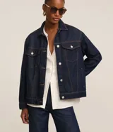 Campera trucker de jeans azul oscuro, corte amplio, con cuello clásico, cierre frontal con botones blancos, mangas largas y bolsillos delanteros con solapa.