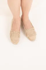 Mocasín color beige de cuero gamuzado con flecos y lazo con herrajes metálicos dorados en la capellada. Interior forrado en cuero y suela de goma flexible.