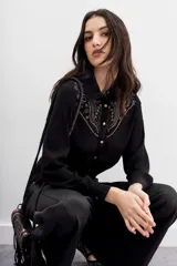 Camisa negra de mangas largas con cuello camisero y botones metálicos. Presenta tachas termofijadas en el frente y espalda.