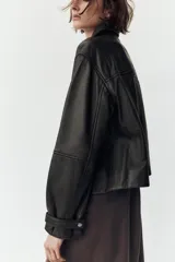 Cazadora negra de cuero con cuello solapa, manga larga con trabilla y bolsillos de parche con solapa en el delantero. Cierre frontal con cremallera y botones a presión ocultos por solapa.