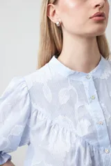Camisa de algodón con fondo azul claro a rayas finas y estampa floral blanca en relieve. Presenta cuello cerrado, cartera de botones frontal y mangas cortas con volumen y puño elastizado. Posee frunces en el frente que aportan caída.