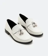 Zapato mocasín de color blanco con textura de piel de cocodrilo y detalle de borla frontal. Posee punta redonda, tacón plano y suela en contraste de color negro.