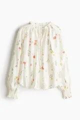 Blusa oversized de viscosa color crema con estampado floral. Tiene cuello corto con volado y abertura en pico con tiras finas. Las mangas son raglán largas con elástico fino en los puños y el bajo es redondeado.
