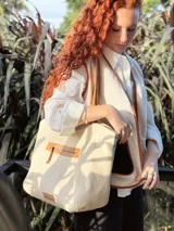 Bolso tipo tote color crema, con detalles en cuero color marrón. Tiene un bolsillo con cierre en la parte superior y un parche de cuero con el logo de la marca en la parte inferior.