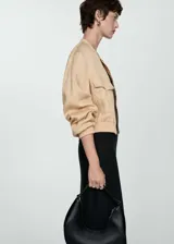 Chaqueta bomber color beige con cierre frontal, cuello redondo, mangas largas con puños elásticos y dos bolsillos delanteros con solapa.