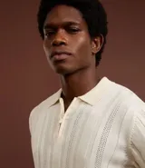 Camisa de punto color crema, con cuello polo y cierre frontal parcial con cremallera, de manga corta y diseño texturizado con perforaciones verticales.