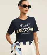 Remera azul marino de algodón con cuello redondo y mangas cortas. Estampa frontal con la palabra "Merci" en color crema, dos corazones entrelazados con detalles de perlas y la frase "mon amour" en cursiva.