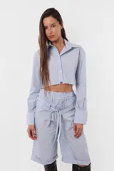 Camisa celeste crop confeccionada en poplín de algodón, con terminación al corte para que desfleque. Acceso a la prenda mediante cartera de botones, cuello y puño reforzado. En canesú de espalda detalle de bordado aplique al corte de logo TW.