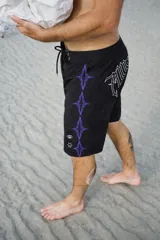 Short de baño negro con cordón de ajuste blanco en la cintura. Presenta un estampado vertical de figuras abstractas en color violeta y un logo estilizado en blanco en el lateral derecho. En el ruedo inferior izquierdo tiene un pequeño logo circular y una estrella de cinco puntas.