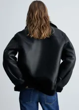Chaqueta biker negra de efecto piel con cuello de borreguito, cierre de cremallera asimétrico, bolsillos laterales con cremallera y terminaciones de borreguito.