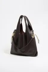 Bolso de mano tipo shopper de cuero color marrón oscuro, con estructura blanda y asa extraíble. Incluye logo metálico de la marca Bimba y Lola, acabados metálicos, bolsillo interior e interior con etiqueta de tejido.