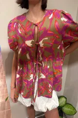 Blusa de calce holgado con estampado floral en tonos marrones, fucsias, naranjas y blancos. Escote en V con doble lazo ajustable y mangas 3/4 con volumen.