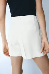 Short blanco de corte sastrero con cierre y botón, bolsillos al bies y bolsillo trasero decorativo. Piernas con pinzas y pliegues adelante y atrás.