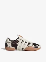 Zapatillas Adidas Taekwondo Mei sin cordones, con capellada de cuero vacuno con estampado animal print de vaca, las tres tiras características de la marca en cuero blanco y suela de goma color caramelo.
