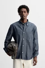 Camisa azul de algodón con textura chambray, corte regular fit, cuello abotonado, manga larga con puños abotonados, bolsillo de parche en el pecho y cierre frontal con botones.
