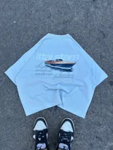 Remera amarilla de corte holgado con estampado trasero de un yate y texto con nombres de ciudades.